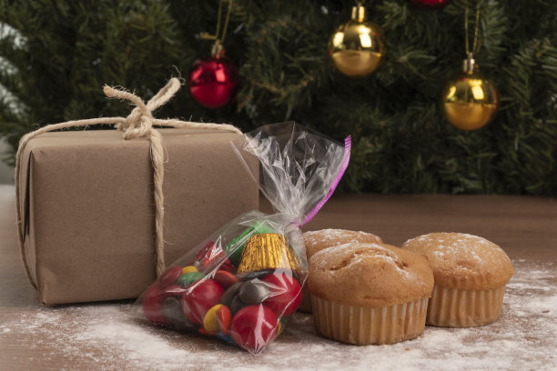 圣诞庆祝与礼品包装旁边的纸杯蛋糕和一个盖子与糖果和巧克力，食物与圣诞树的背景，工作室图片下载