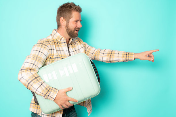 年轻的微笑随意的男人拿着手提箱指着手指面孤立的绿色背景。图片下载