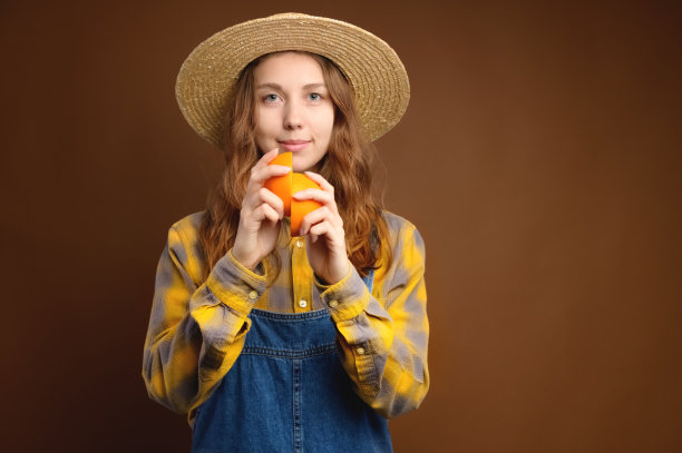 戴着草帽的迷人的白人年轻女子手里拿着切开的橘子。工作室肖像在棕色背景。看着摄像机图片下载