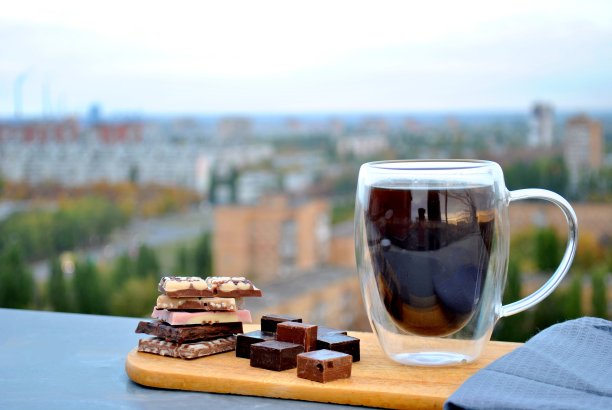 一个透明的杯子里装着黑色的热咖啡和几块不同的巧克力，在晨曦中朦胧的城市背景上。图片下载