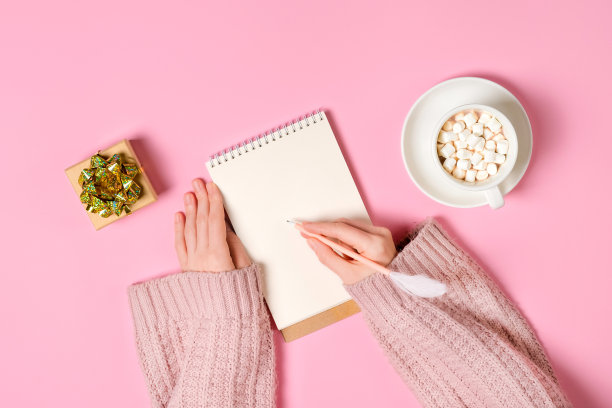 记事本，金箔，和一杯热巧克力与棉花糖和女性的手写孤立的粉红色背景。新年，圣诞节或节日愿望清单横幅与复制空间图片下载