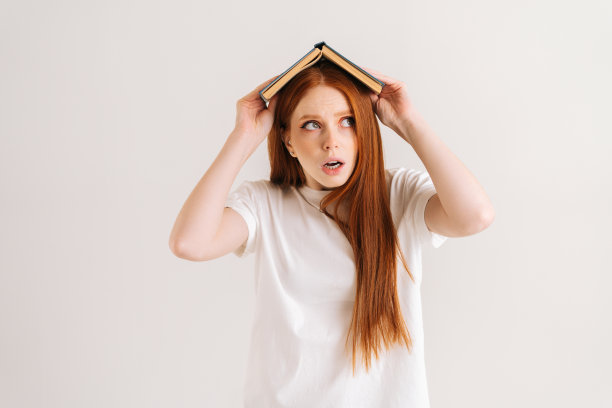 工作室肖像紧张的年轻学生妇女盖着头与书看别处，站在白色孤立的背景。图片下载
