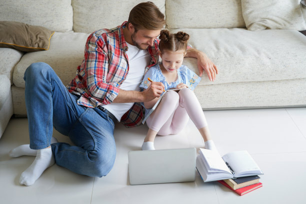 微笑的父亲和女儿坐在客厅的地板上，带着笔记本电脑，上课。图片下载