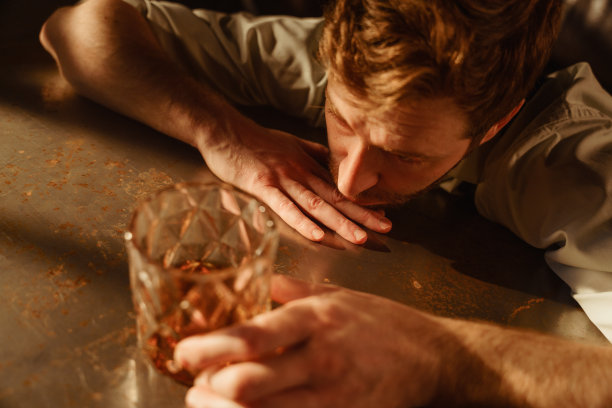 餐桌上一个喝着威士忌的醉汉的大气肖像。一张从桌子上方拍摄的照片。那人把头靠在桌子上，右手托着下巴，左手拿着一杯威士忌。图片下载