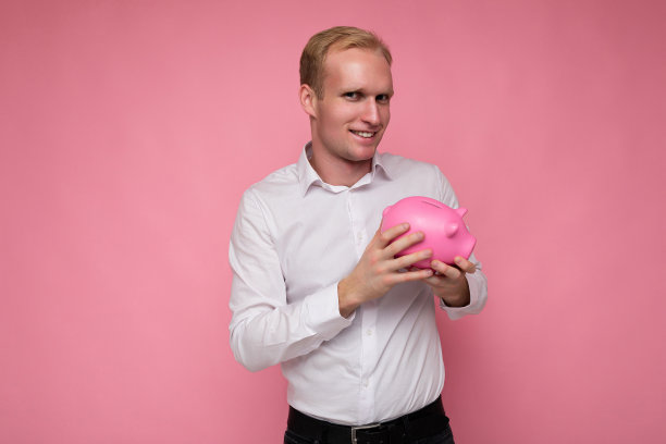肖像照片的快乐积极微笑年轻英俊的金发男人真诚的情绪穿着休闲的白色衬衫孤立在粉红色的背景与复制空间和拿着粉红色的小猪盒子的钱。银行的概念图片下载