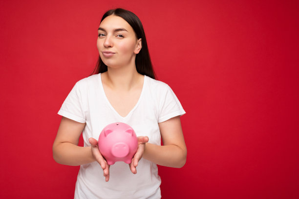 照片肖像积极自信的年轻美丽漂亮的女人与长黑发真诚的情绪穿着休闲的白色t恤孤立在红色的背景与自由的空间和持有粉红色的小猪盒子。储钱罐的概念图片下载