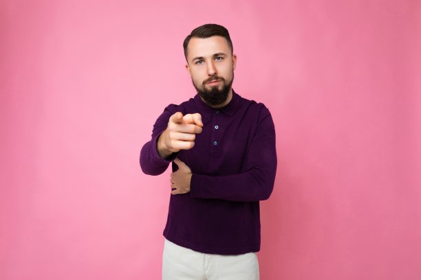 照片拍摄的英俊自信和好看的年轻深色胡子男子穿着休闲和时尚的紫色长袖摆姿势孤立在粉红色的背景，空白的文字空间，并对着相机手指图片下载