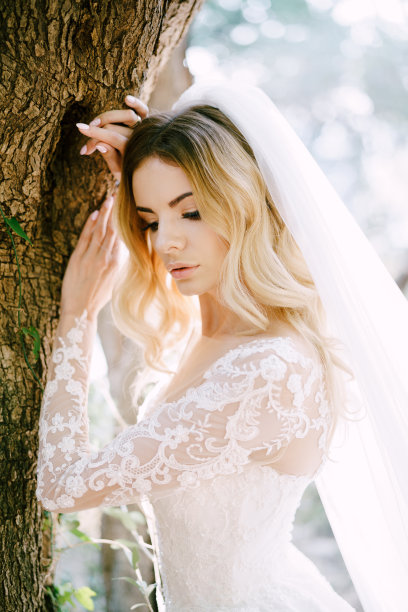 新娘身穿白色婚纱，头戴面纱，倚在树干上图片下载