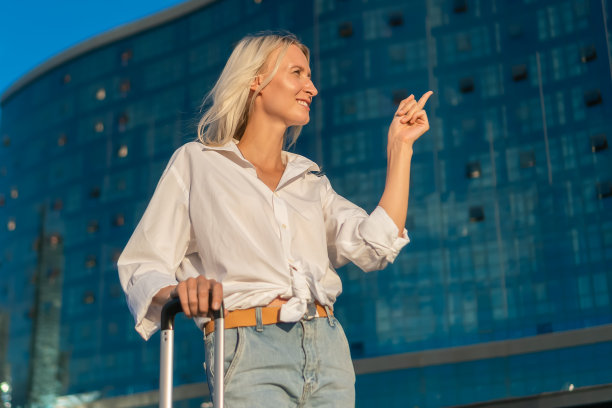 年轻快乐的金发女人穿着白衬衫和牛仔裤，另一只手拿着手提箱，手指指向一边，在阳光下的一个玻璃旅馆的背景下，复制空间图片下载