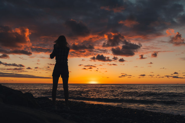 一个年轻苗条的女人的剪影，站在海边，用手机拍摄美丽、多彩的、神奇的日落图片下载