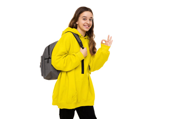 漂亮的、可爱的、微笑的、积极的棕发少女身穿时尚的黄色运动帽衫，背着灰色背包，做出OK手势，在白色背景上隔离，有文字的自由空间照片图片下载