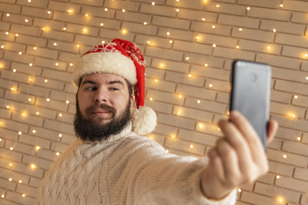 年轻的成年大胡子男人圣诞节自拍肖像在装饰室内白墙黄花环无聚焦照明背景图片下载