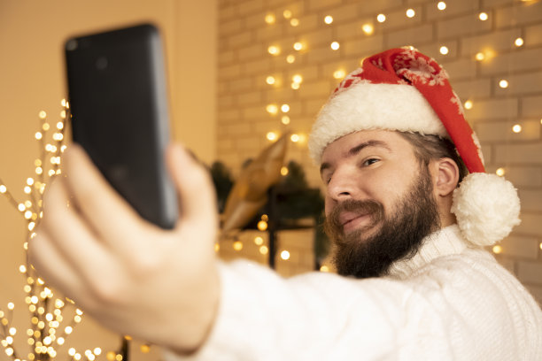 圣诞自拍，戴着红帽的大胡子男人在冬季假期的孤独概念房间室内环境空间与墙上的花环灯的背景图片下载