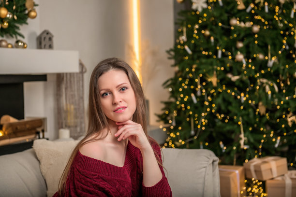 一个年轻漂亮的女人正坐在客厅的沙发上。新年的心情和室内装饰着圣诞树上的花环。图片下载