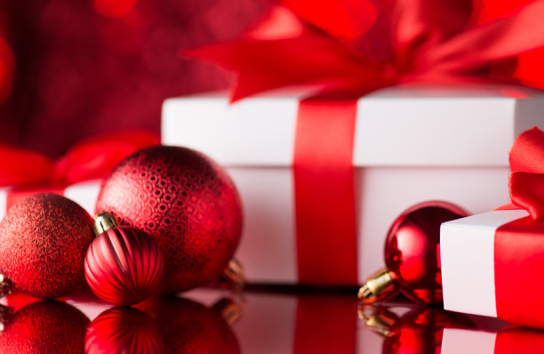红色圣诞树装饰物的特写照片，装饰物球和白色礼品盒，红色丝带蝴蝶结在红焦背景上图片下载