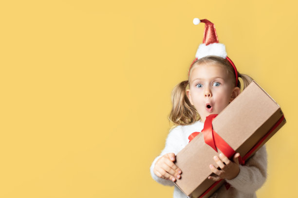 送圣诞礼物。惊讶兴奋的小女孩戴着红色圣诞老人帽摆出孤立的黄色背景抱着牛皮纸纸盒礼物。网上购物，个人产品销售。模拟、空间复制图片下载