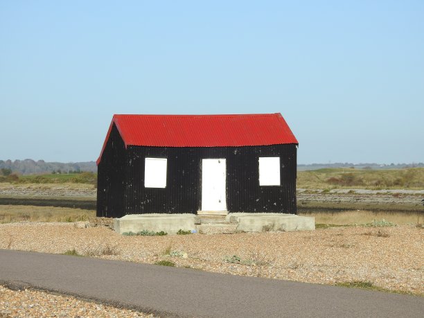 前视图的黑色小屋与红屋顶，白色的门和两个白色的窗户，站在混凝土道路之外，这条道路在黑麦港自然保护区的瓦片区域之间图片下载