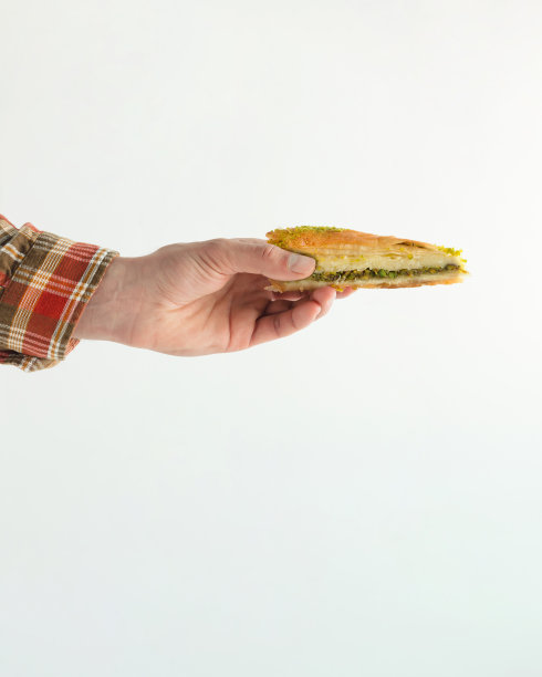 穿着土色格子衬衫的男性手拿着一块果仁饼，上面有开心果。最小的美味食品场景孤立在柔和的白色背景。传统东方烘焙糖果概念。图片下载