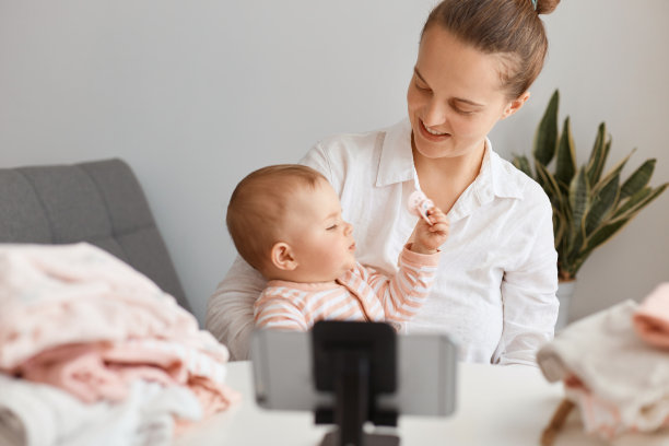 肖像的吸引力积极的年轻成年妇女与婴儿女儿在三脚架前与手机，为她的视频博客拍摄视频，看着她迷人的孩子。图片下载