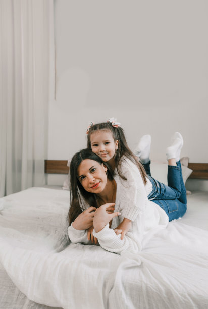 一个年轻的女儿在床上和她年轻的妈妈玩耍，跳在她妈妈的背上，玩得很开心，玩得很开心。他们看着摄像机图片下载