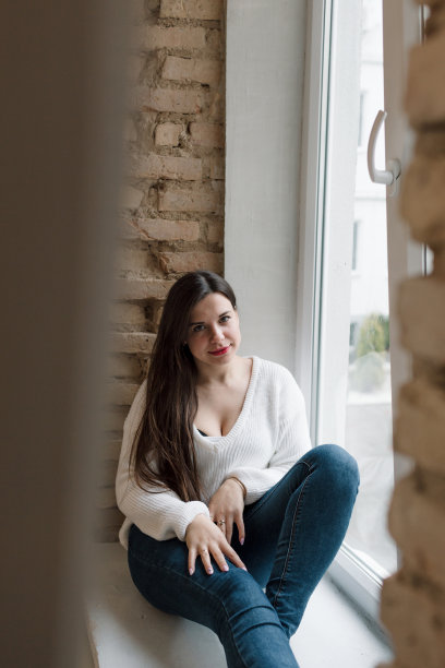 一个穿着白色毛衣和牛仔裤的快乐女孩坐在彩色玻璃窗上，这是一个砖砌的工业阁楼风格的室内。休闲的概念。图片下载