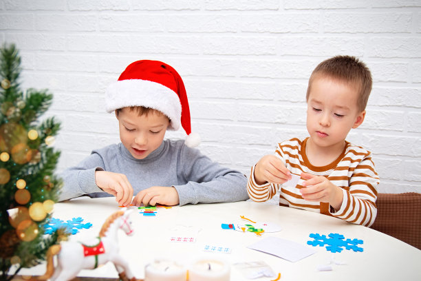 戴着圣诞老人帽子的孩子们坐在桌旁做圣诞工艺品。兄弟们正在为新年准备礼物。学前教育和早期发展图片下载