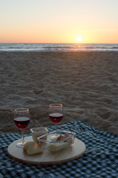 日落时分，在沙滩上野餐，木制托盘上放着两杯红酒、小吃、沙拉三明治和奶酪，还有绿色方格桌布图片下载