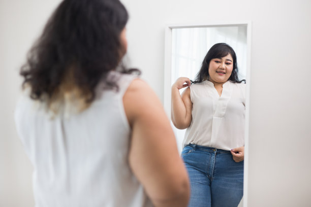 身体正面加尺寸的女性服装。积极的身体加上大小的女人快乐和自豪的自己看着镜子在家里。试着穿合身的衣服。图片下载