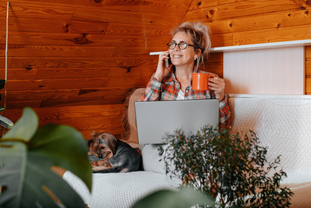 微笑的成年女性自由职业者，打电话，喝咖啡或茶，而工作的笔记本电脑，而坐在家里的狗在早上。快乐女商人远程工作，自由职业者，远程工作图片下载