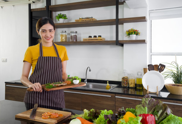 年轻的亚洲家庭主妇穿着围裙微笑着拿着美味的牛排。现代厨房的早晨氛围图片下载