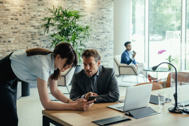 公司业务:两位同事在现代开放式办公室里寻找手机并一起讨论图片下载