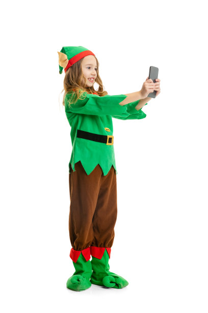 可爱的微笑的小女孩打扮得像有趣的侏儒或小精灵使用电话隔离在白色工作室的背景。冬天，假期，圣诞节的概念图片下载