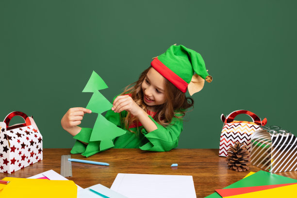 一个快乐的孩子打扮得像有趣的侏儒或小精灵坐在桌旁，在绿色的背景上剪下一棵隔离的纸圣诞树。图片下载