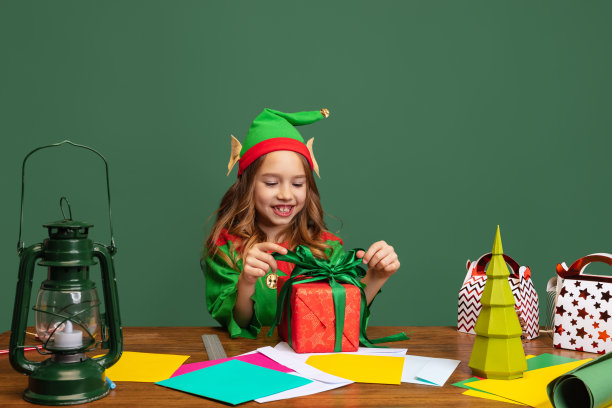 微笑的小女孩，打扮得像有趣的侏儒或小精灵坐在餐桌旁，与节日，圣诞礼物盒孤立在绿色的背景。图片下载