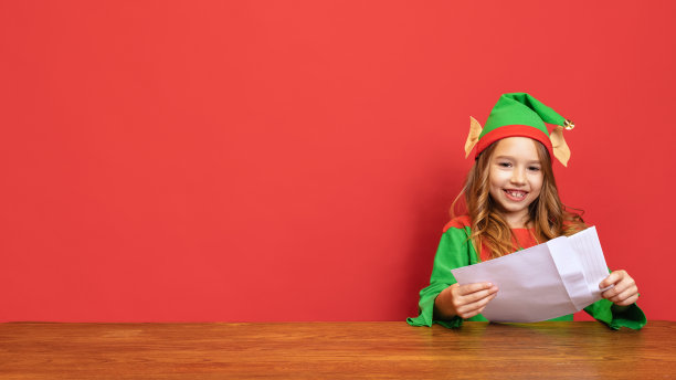 肖像快乐的孩子打扮成有趣的侏儒或小精灵坐在桌旁，写信件给圣诞老人孤立在红色的背景。图片下载