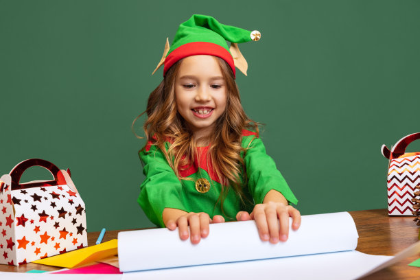肖像快乐的孩子打扮得像有趣的侏儒或小精灵坐在桌旁和写信给圣诞老人孤立的绿色背景。图片下载