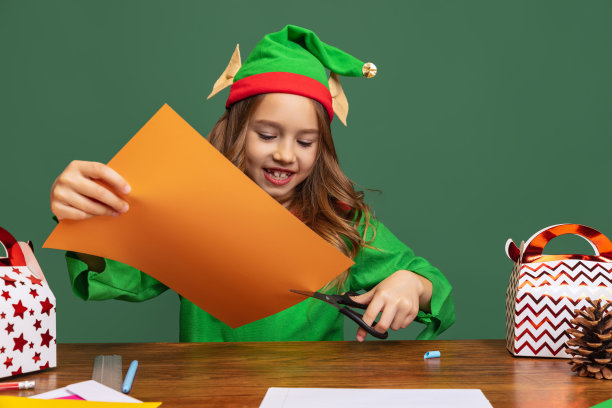 一个可爱的小女孩打扮得像有趣的侏儒或小精灵坐在桌旁，在绿色的背景上做圣诞装饰。图片下载