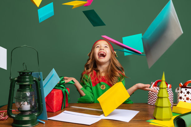 小快乐的孩子，女孩打扮得像有趣的侏儒或小精灵坐在桌旁，帮助圣诞老人阅读圣诞邮件隔离在绿色的背景。图片下载