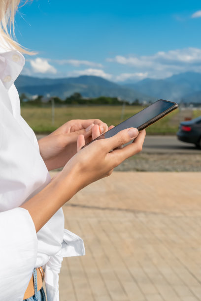 在阳光明媚的日子里，女人的手拿着手机，背景是山脉，汽车特写。垂直图图片下载
