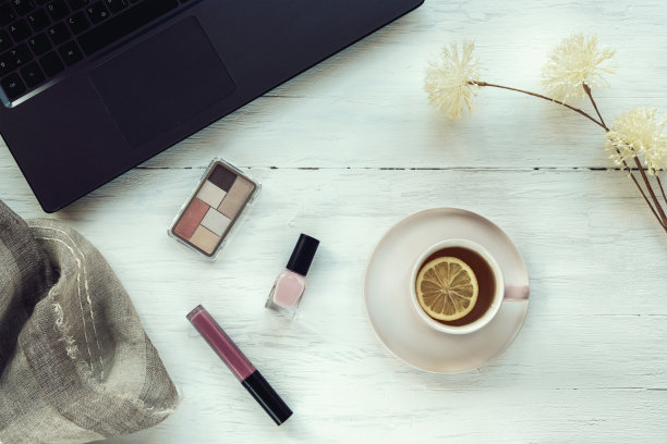 白色木桌上放着笔记本电脑、一杯茶、化妆品和女性配饰。俯视图，平铺图片下载
