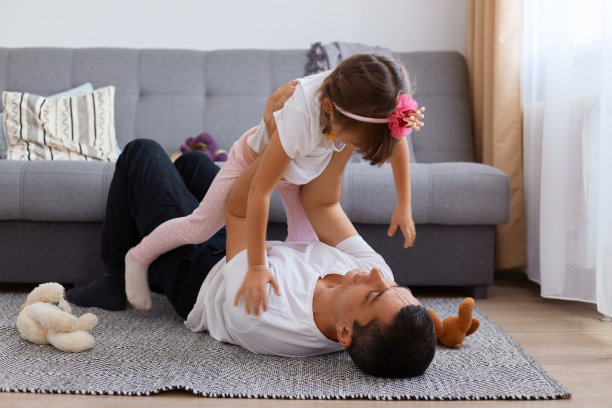 在室内拍摄的照片中，快乐的迷人的年轻成年男子躺在沙发附近的地板上，和他迷人的女儿玩耍，把他的孩子抱起来，穿着白色t恤和裤子。图片下载