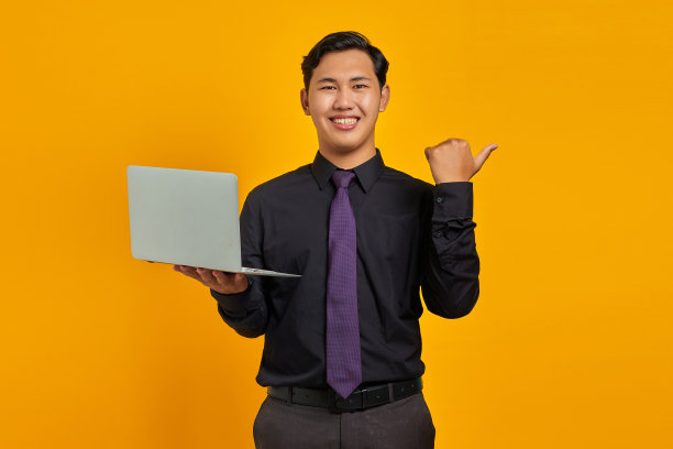 微笑的亚洲商人拿着笔记本电脑，大拇指指向黄色背景上的复制空间图片下载