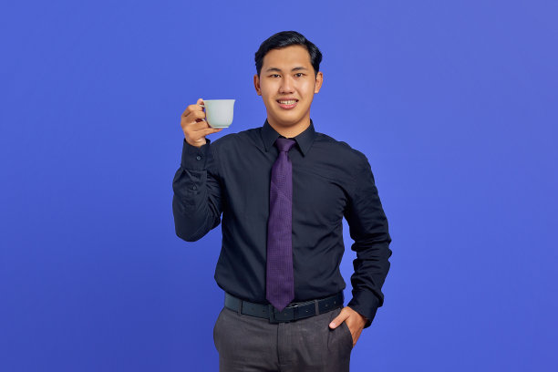 英俊的年轻商人在相机微笑，手在口袋里，拿着杯子在紫色的背景上图片下载