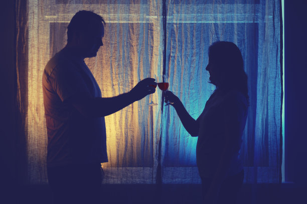 一个男人和一个女人在夜窗边喝酒的剪影。已婚夫妇有饮酒问题在晚上的家庭客厅灯光图片下载