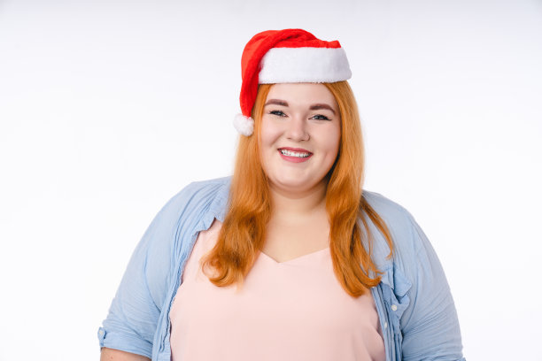 丰满的大尺码年轻美丽的女人笑在圣诞老人的红帽子看着相机准备圣诞节新年孤立在白色的背景图片下载