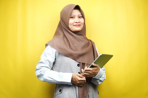 美丽的年轻亚洲穆斯林妇女微笑，兴奋和愉快的拿着平板，孤立在黄色背景图片下载