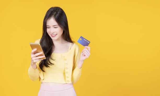 金融支付网上购物和转账概念。愉快的微笑年轻女子拿着智能手机和网上银行的模型信用卡，而站在孤立的黄色背景。图片下载
