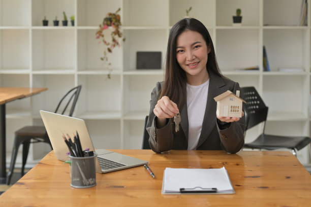 照片上，一个漂亮的女人拿着房子模型，在木制办公桌前展示钥匙。图片下载