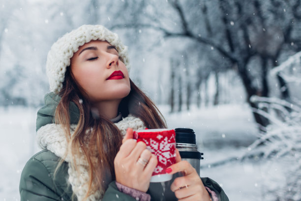 在下雪的冬季公园里手持保温瓶喝热茶的女人。杯子穿着红色针织圣诞外套图片下载