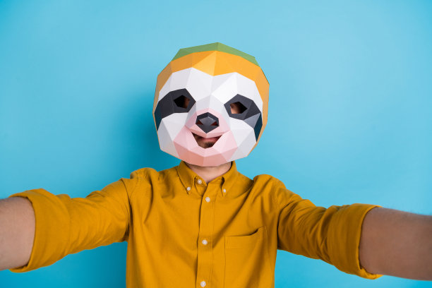 照片的博客树懒男子拿着相机自拍戴着多边形面具黄色衬衫孤立的蓝色背景图片下载